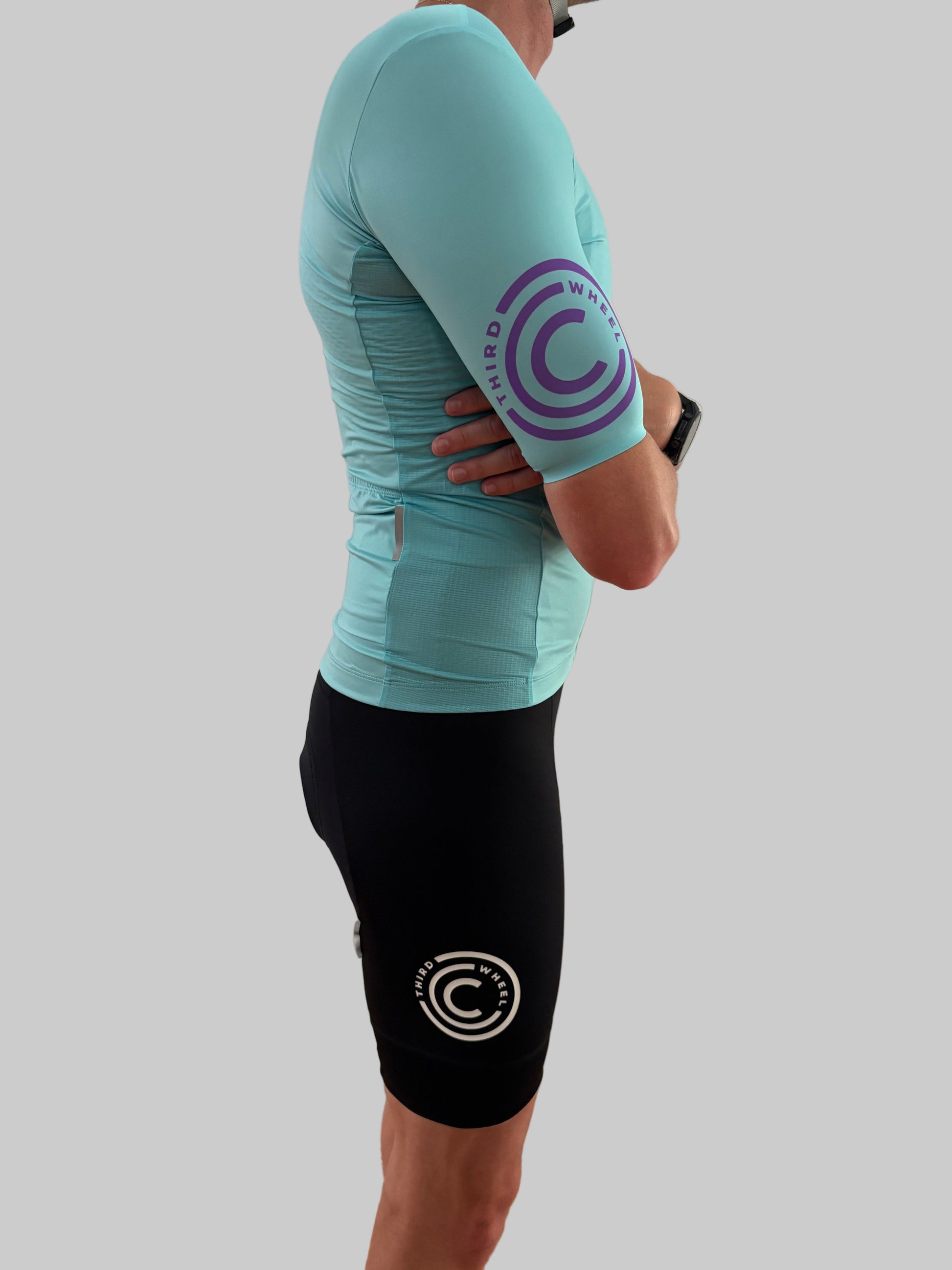 Person wearing a turquoise cycling jersey and black shorts with a logo on a gray background