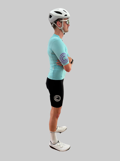 Person wearing a turquoise cycling jersey and black shorts with a logo on a gray background